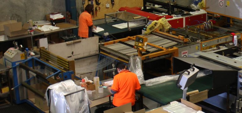 2 Employees In Orange Uniform Working With Heavy Plastic Film Machinery